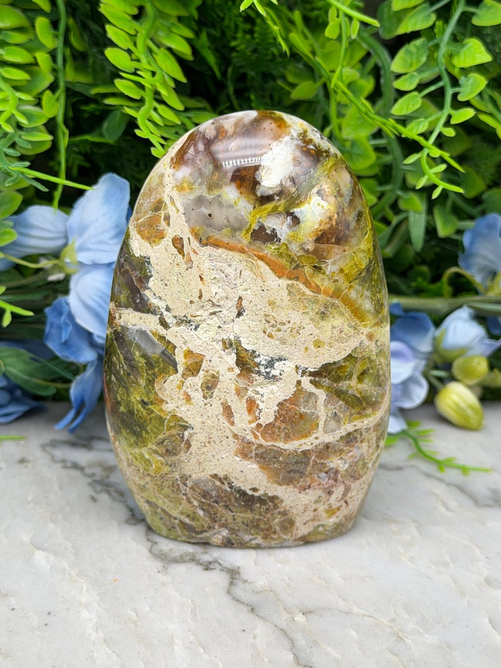 a large, colorful jadeite stone with a unique pattern of green, brown, and yellow hues, set against a backdrop of lush greenery and blue flowers.