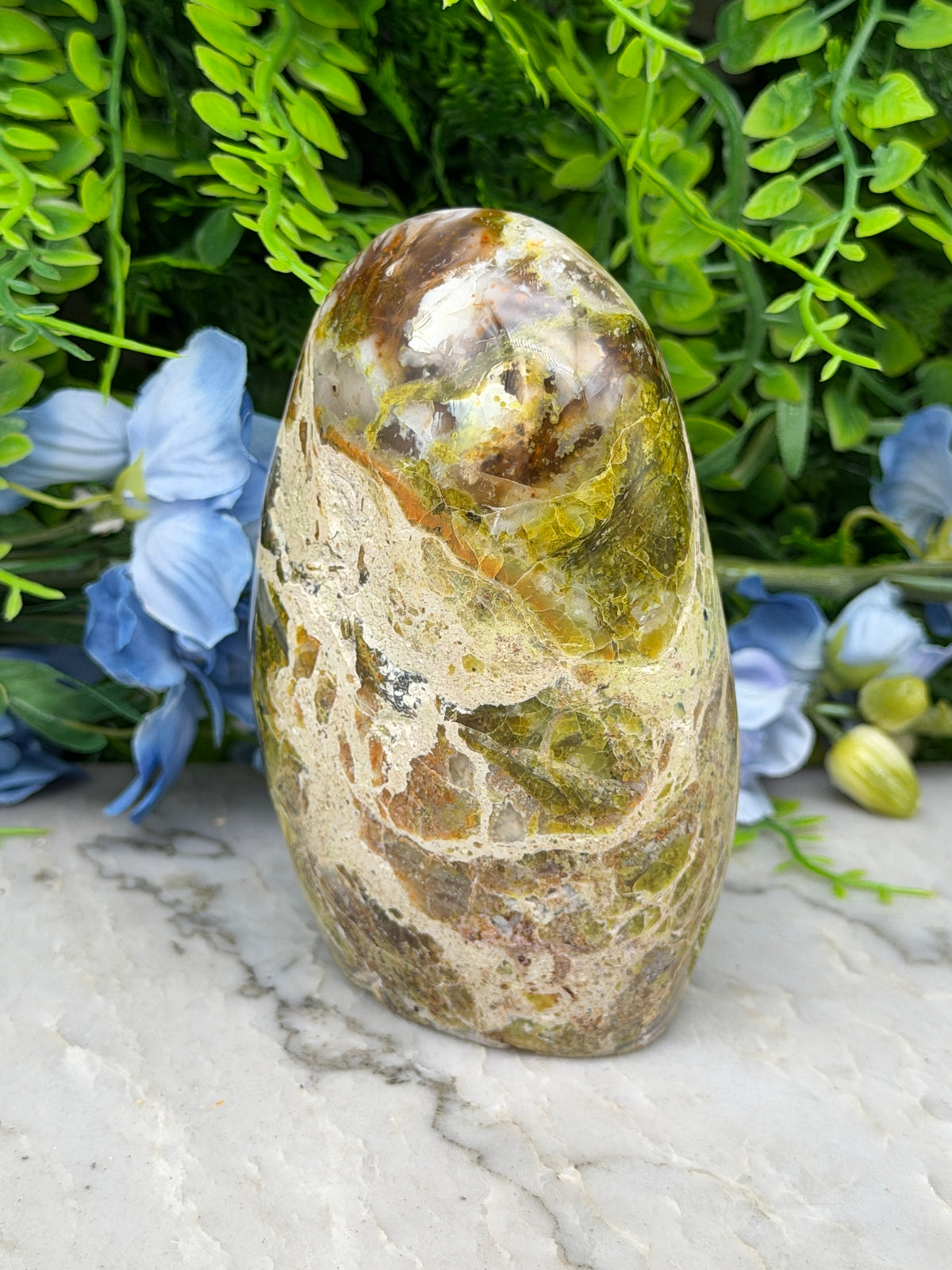 a large, irregularly shaped stone with a mix of green, brown, and yellow colors, set against a backdrop of lush greenery and blue flowers.