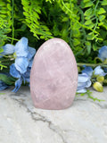 A pink amethyst crystal rests on a marble surface, surrounded by blue flowers and green foliage.
