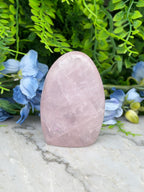 A pink amethyst crystal rests on a marble surface, surrounded by blue flowers and green foliage.