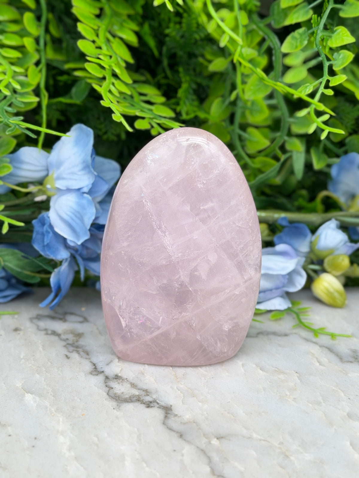 A pink amethyst crystal rests on a marble surface, surrounded by blue flowers and green foliage.