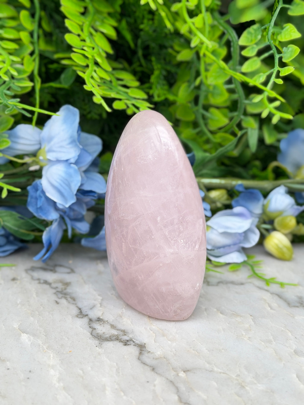 A pink gemstone is placed on a marble surface, surrounded by blue flowers and green foliage.