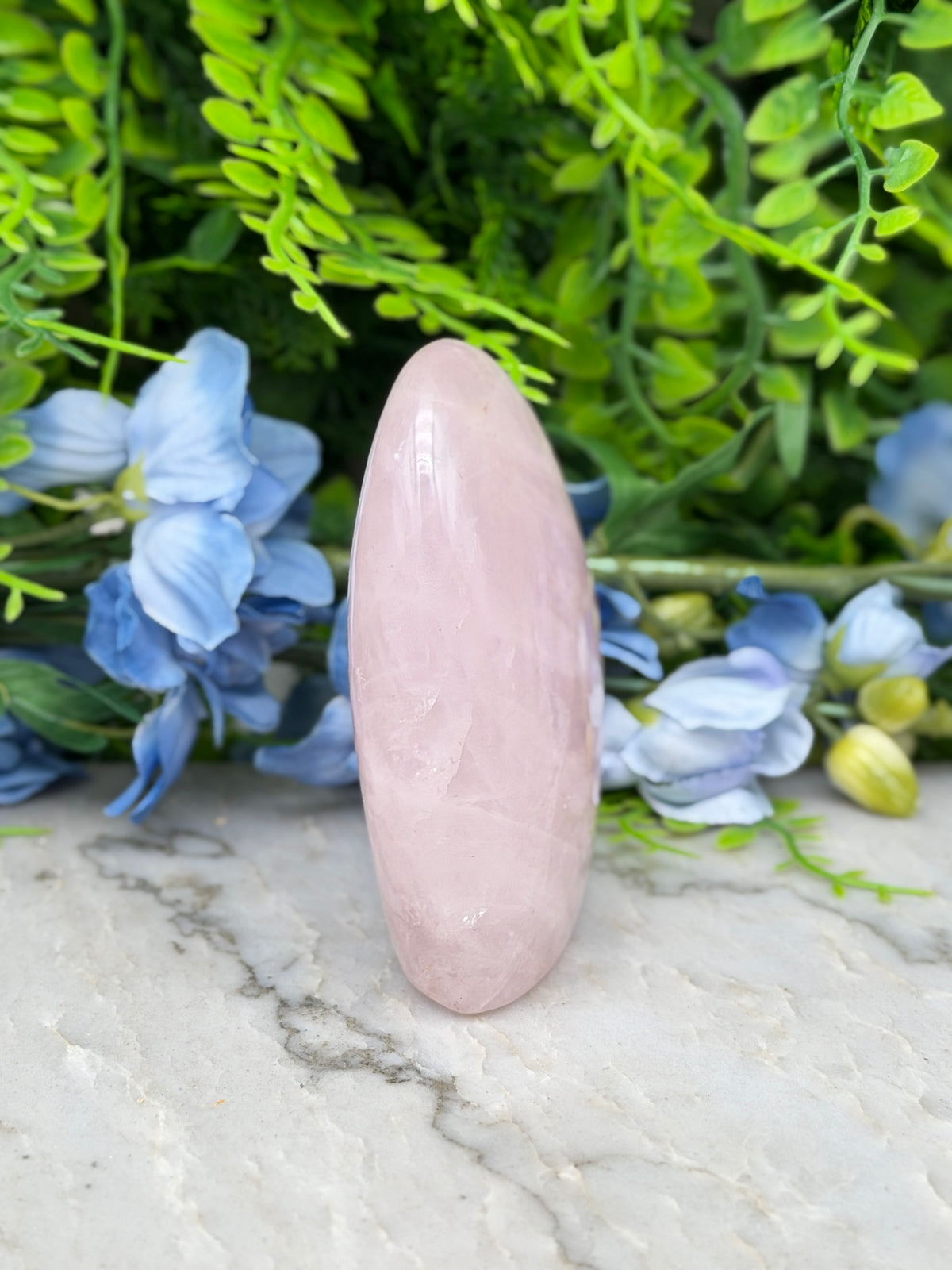 A pink gemstone is placed on a marble surface, surrounded by blue flowers and green foliage.