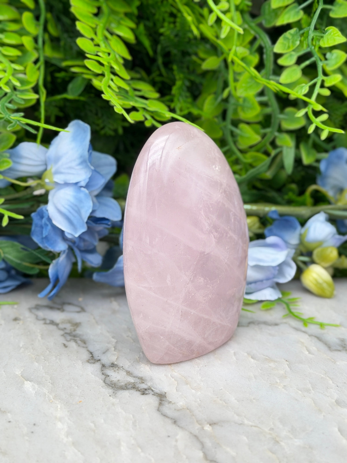 A pink amethyst crystal is placed on a marble surface, surrounded by blue flowers and green foliage.