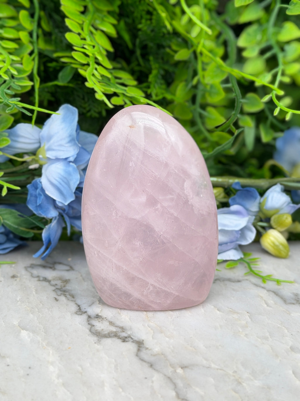 A pink amethyst crystal rests on a marble surface, surrounded by lush green foliage and delicate blue flowers.