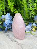 A pink gemstone is placed on a marble surface, surrounded by blue flowers and green foliage.