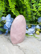 A pink gemstone is placed on a marble surface, surrounded by blue flowers and green foliage.