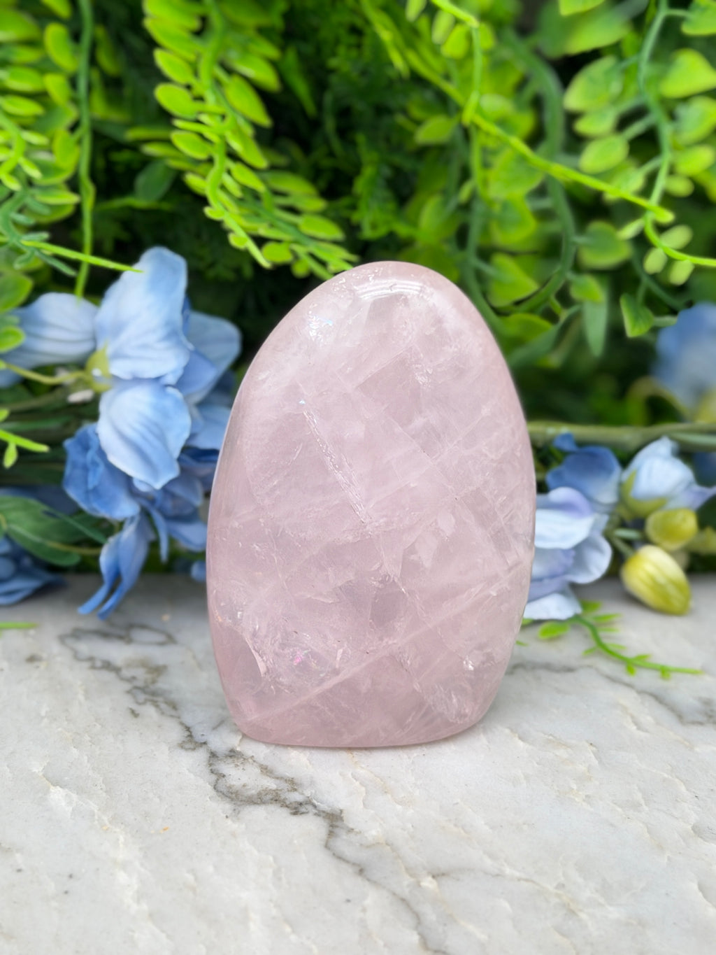 A pink crystal rests on a marble surface, surrounded by lush green foliage and blue flowers.