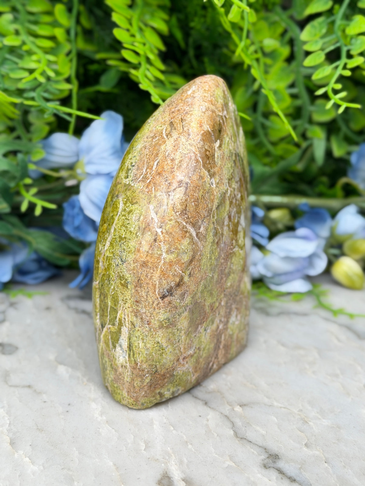 a natural stone with a unique, irregular shape, surrounded by lush green foliage and blue flowers.