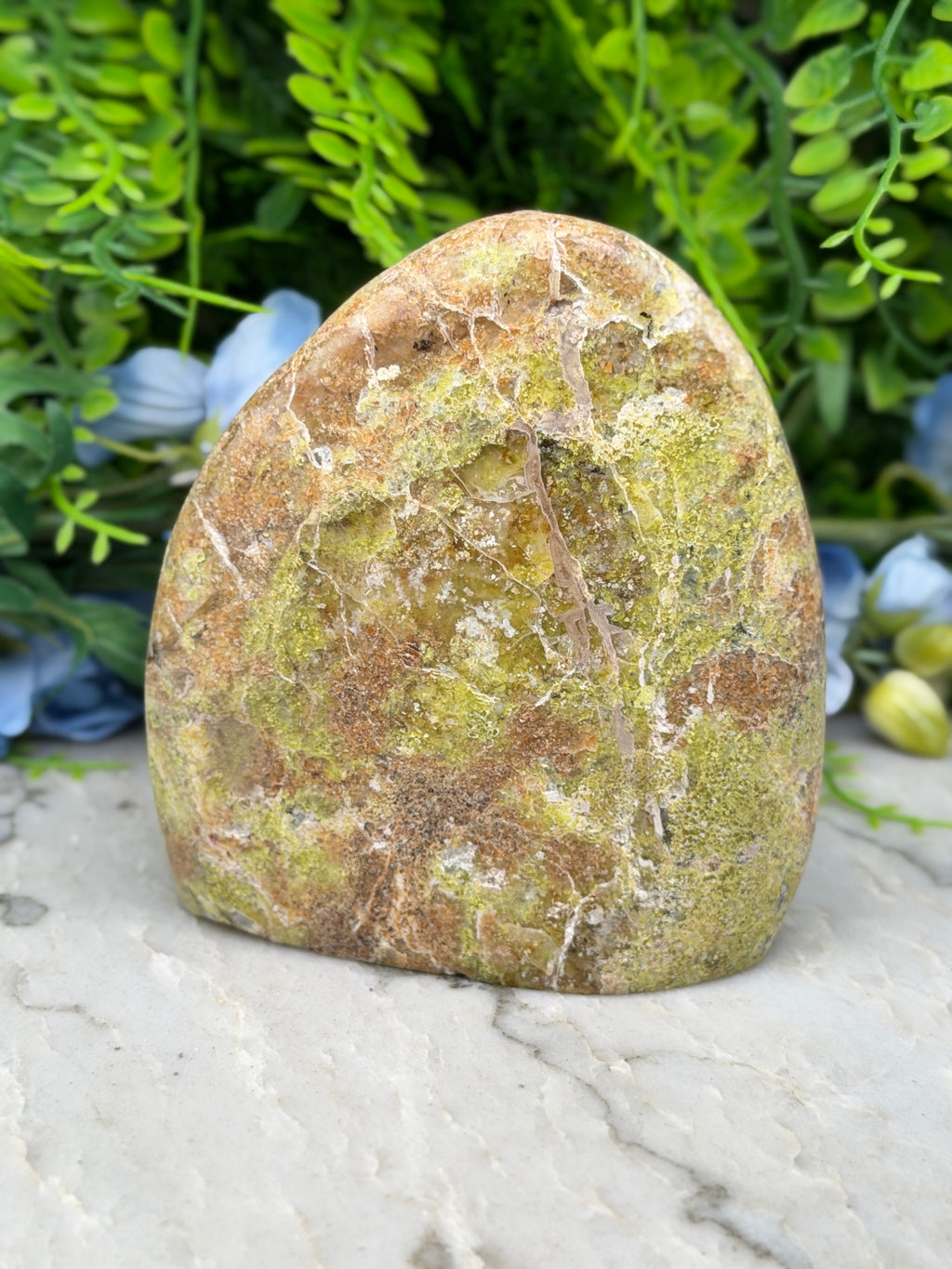 a large, irregularly shaped rock with a mix of green and brown hues, set against a backdrop of lush greenery and flowers.