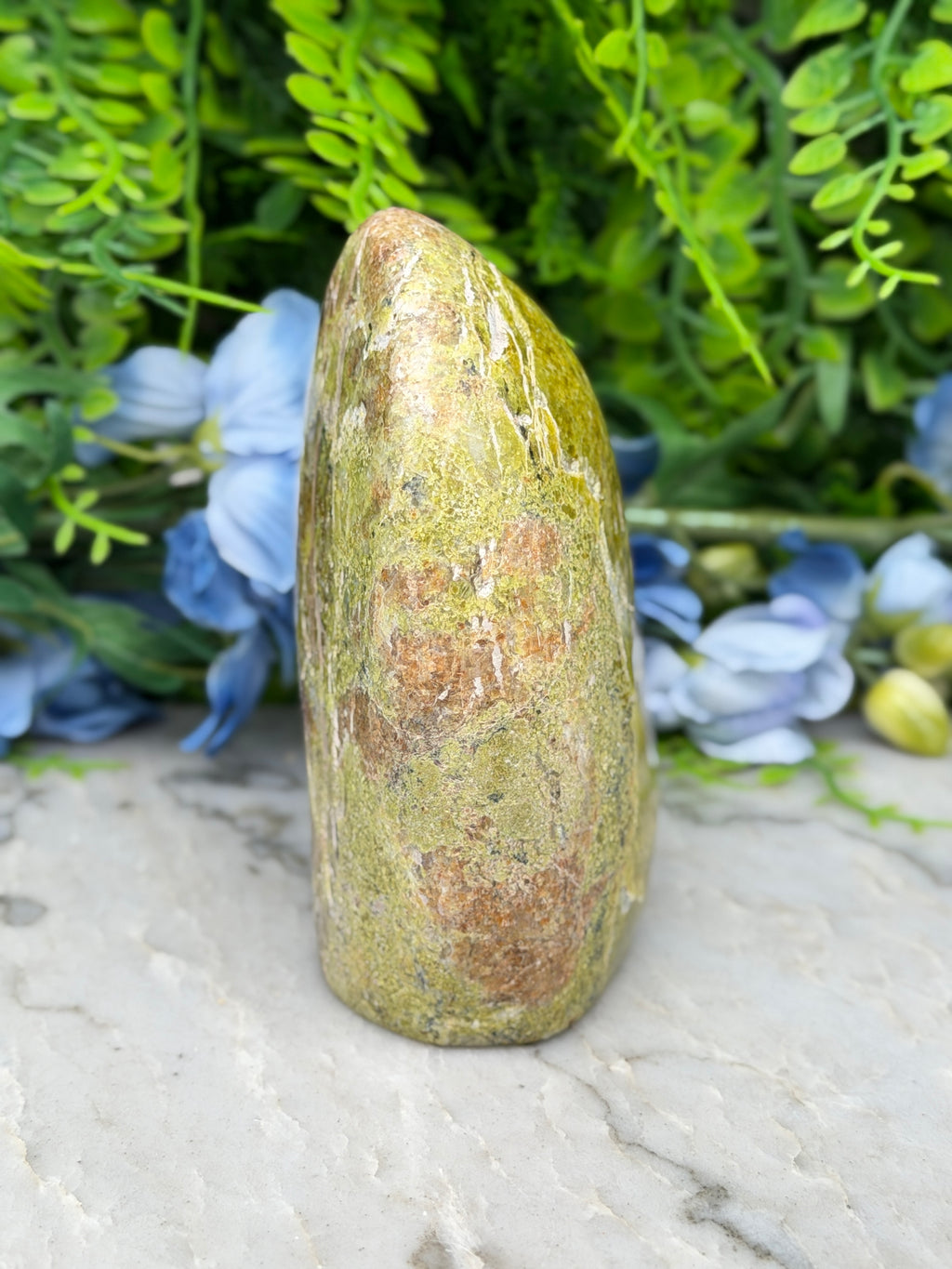 a tall, pointed rock with a greenish-brown hue, set against a backdrop of lush, green foliage and blue flowers.