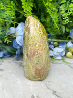 a tall, pointed rock with a greenish-brown hue, set against a backdrop of lush, green foliage and blue flowers.