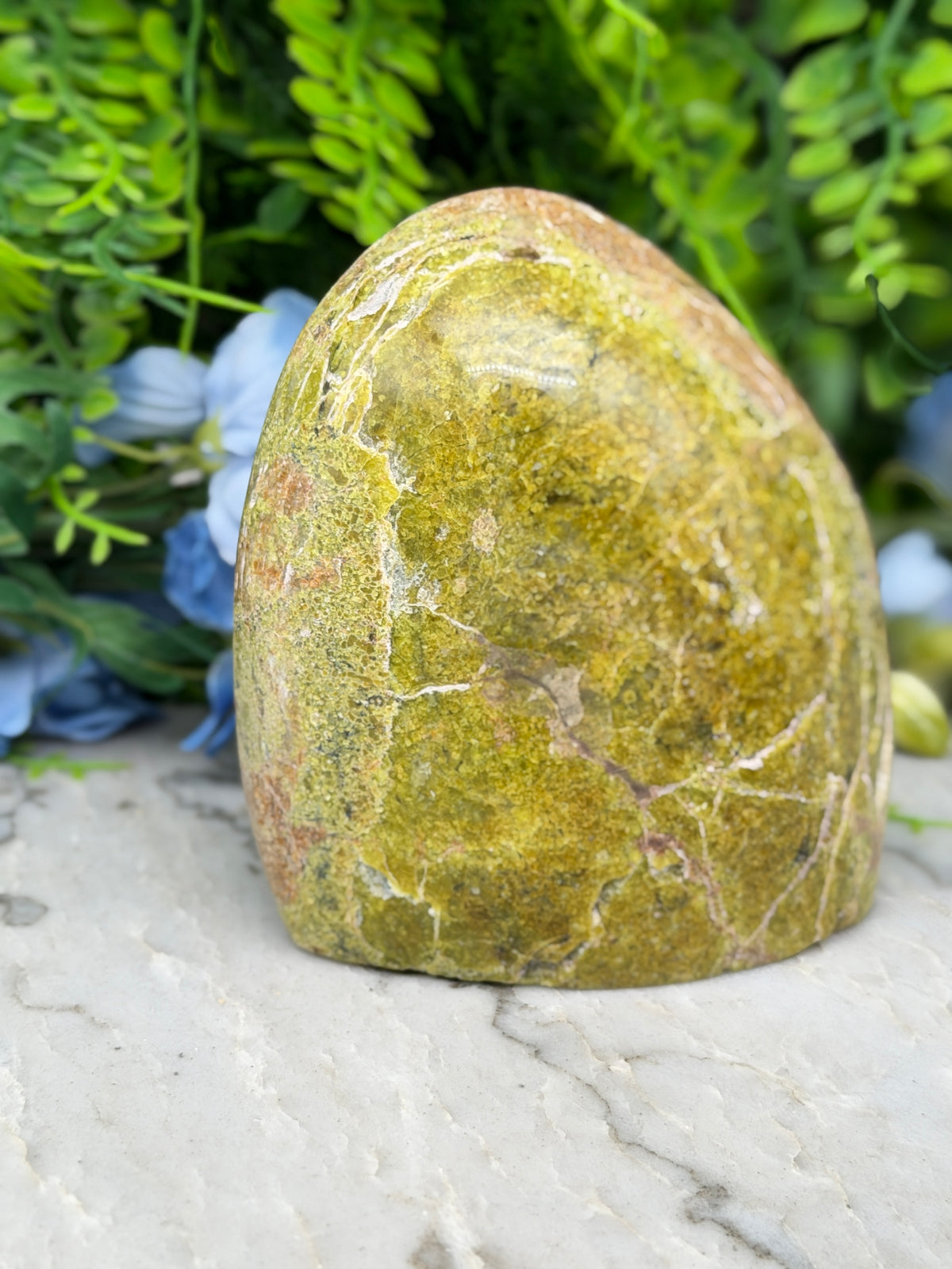 A large, oval-shaped, yellowish rock with a rough, textured surface sits on a white marble surface, surrounded by lush green foliage.
