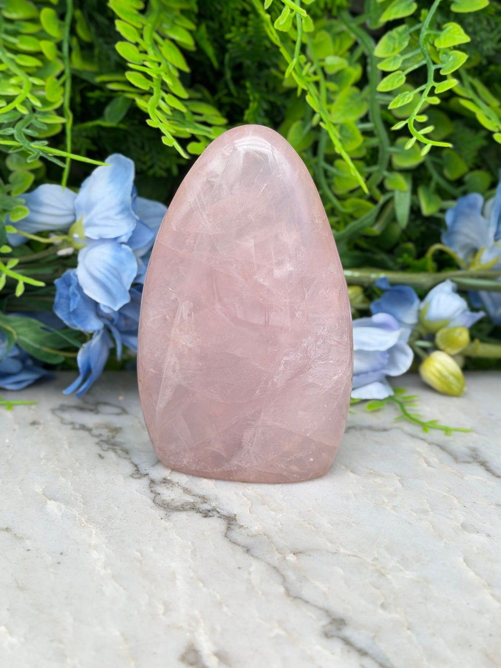 A pink gemstone is placed on a marble surface, surrounded by blue flowers and green foliage.