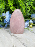 A pink gemstone is placed on a marble surface, surrounded by blue flowers and green foliage.