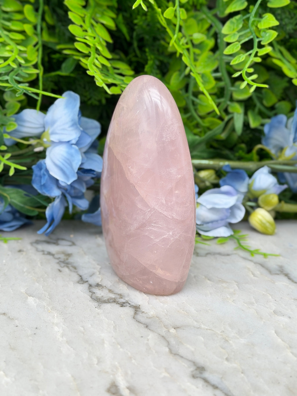 A pink gemstone is placed on a marble surface, surrounded by blue flowers and green foliage.