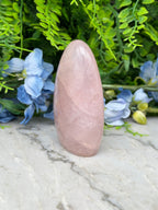 A pink gemstone is placed on a marble surface, surrounded by blue flowers and green foliage.