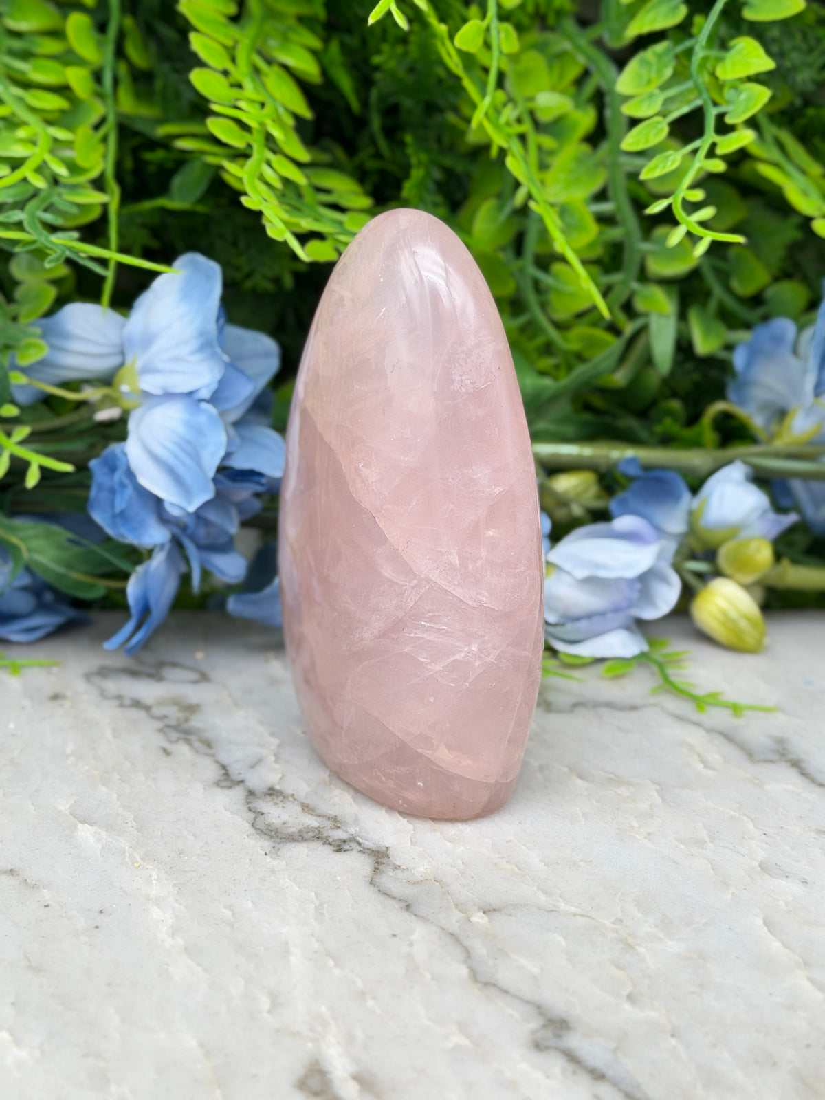 A pink gemstone is placed on a marble surface, surrounded by blue flowers and green foliage.