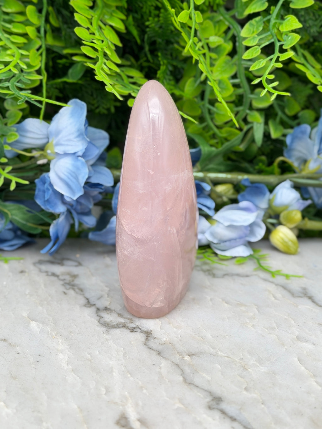 A pink crystal sits on a marble surface, surrounded by blue flowers and green foliage.