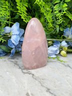 A pink crystal sits on a marble surface, surrounded by blue flowers and green foliage.