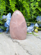 A pink gemstone sits on a marble surface, surrounded by lush green foliage and blue flowers.