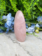 A pink gemstone is placed on a marble surface, surrounded by blue flowers and green foliage.