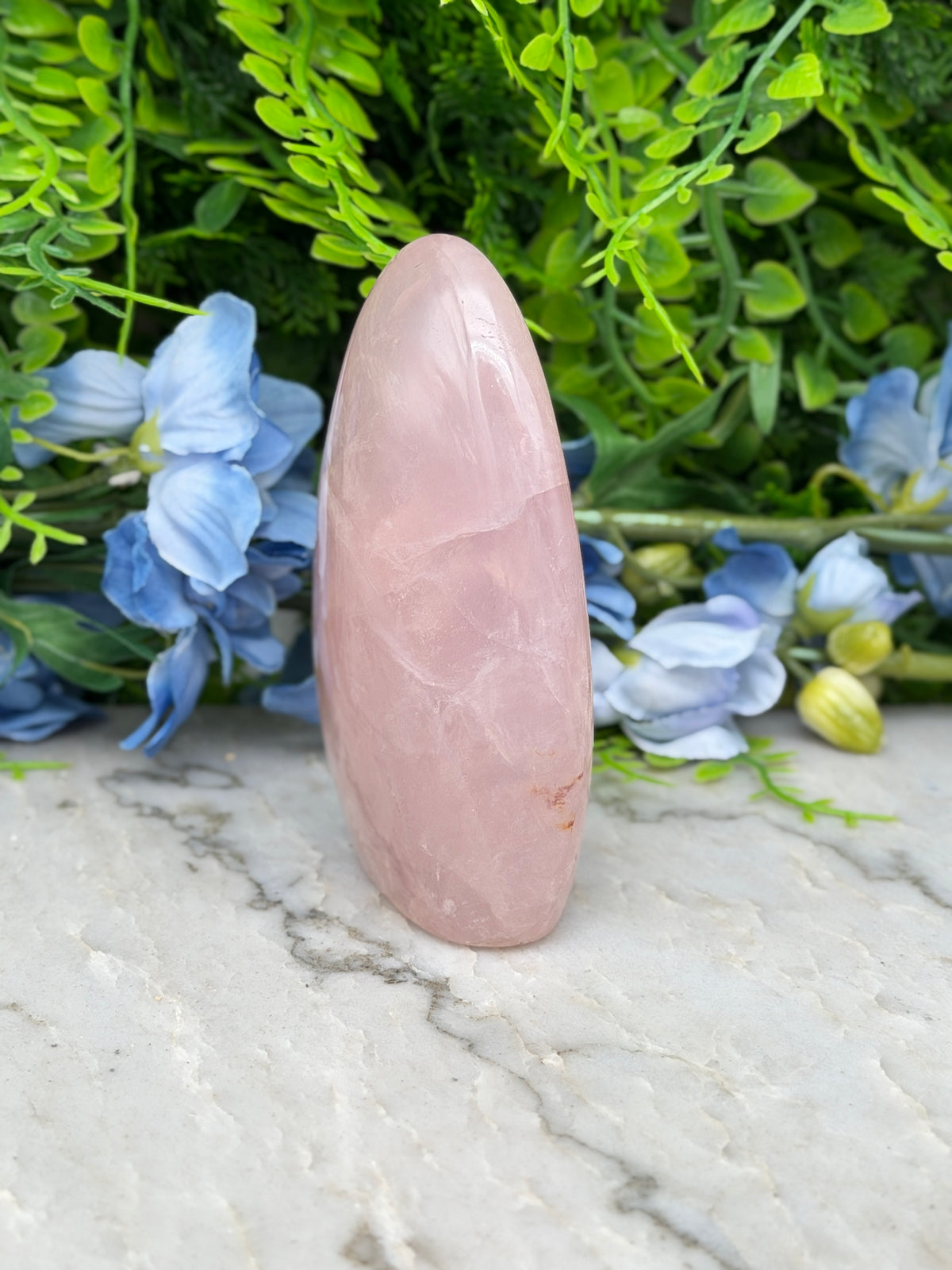 A pink gemstone is placed on a marble surface, surrounded by blue flowers and green foliage.