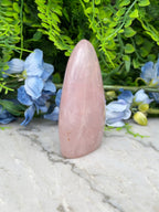 A pink gemstone is placed on a marble surface, surrounded by blue flowers and green foliage.