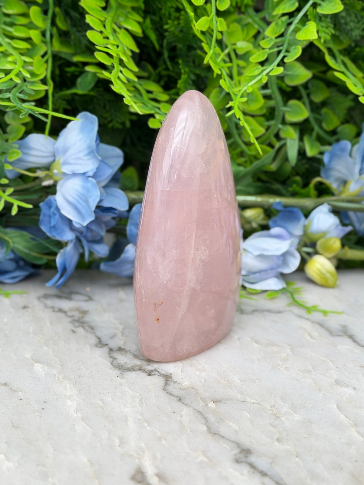A pink gemstone is placed on a marble surface, surrounded by blue flowers and green foliage.