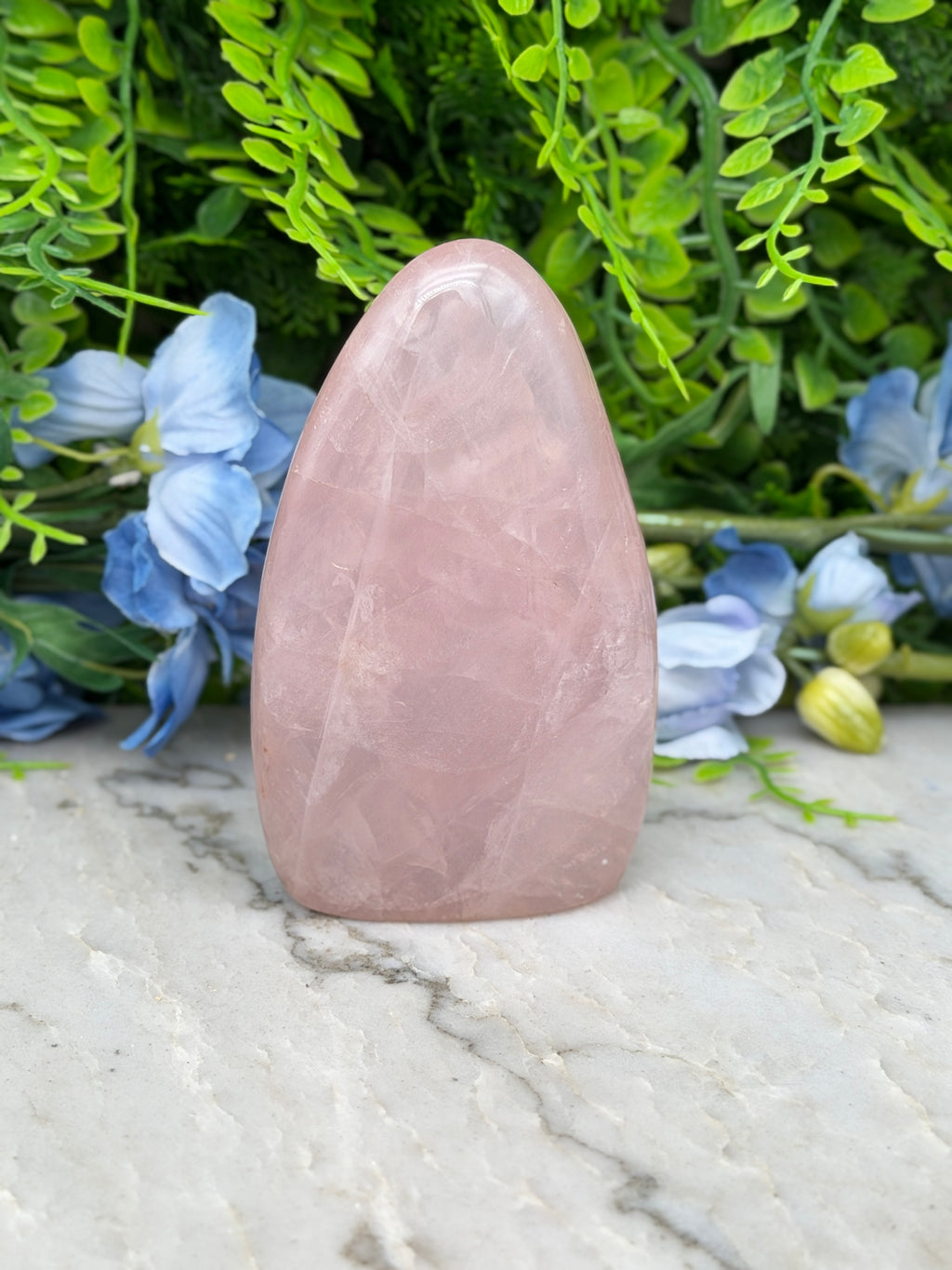 A pink gemstone is placed on a marble surface, surrounded by blue flowers and green foliage.