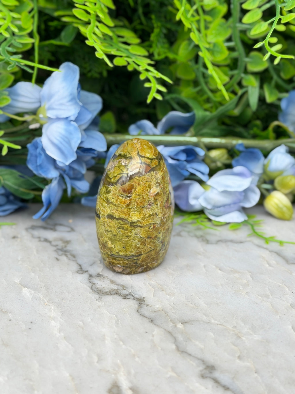 a yellowish-brown stone or rock sitting on a marble surface, surrounded by blue flowers and greenery.
