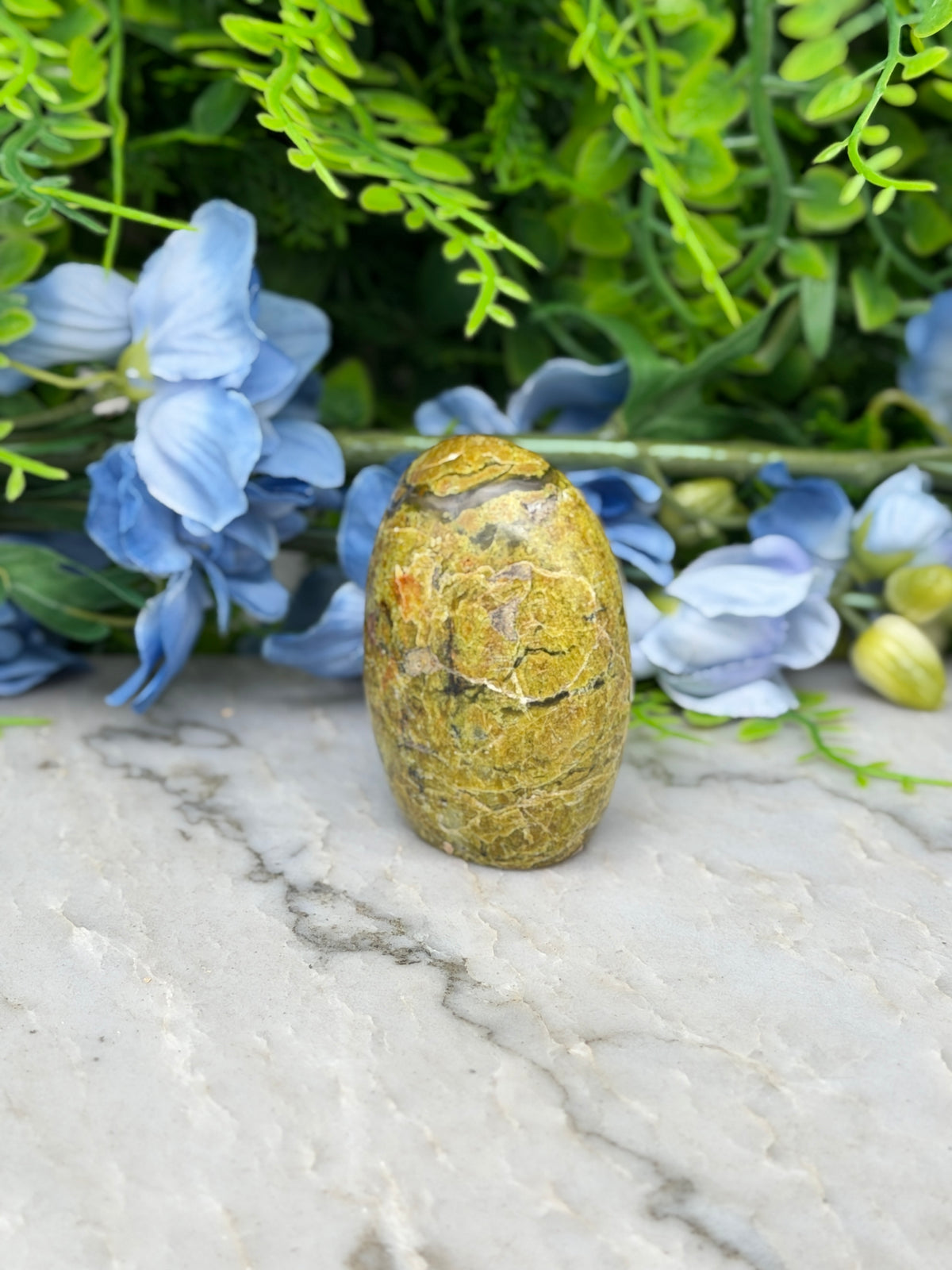 a yellowish-brown stone or rock, possibly a fossil, placed on a marble surface surrounded by blue flowers and greenery.