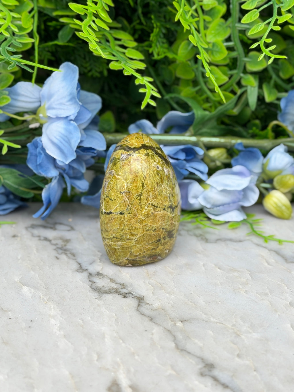 a yellowish-brown stone or rock placed on a marble surface, surrounded by blue flowers and green foliage.