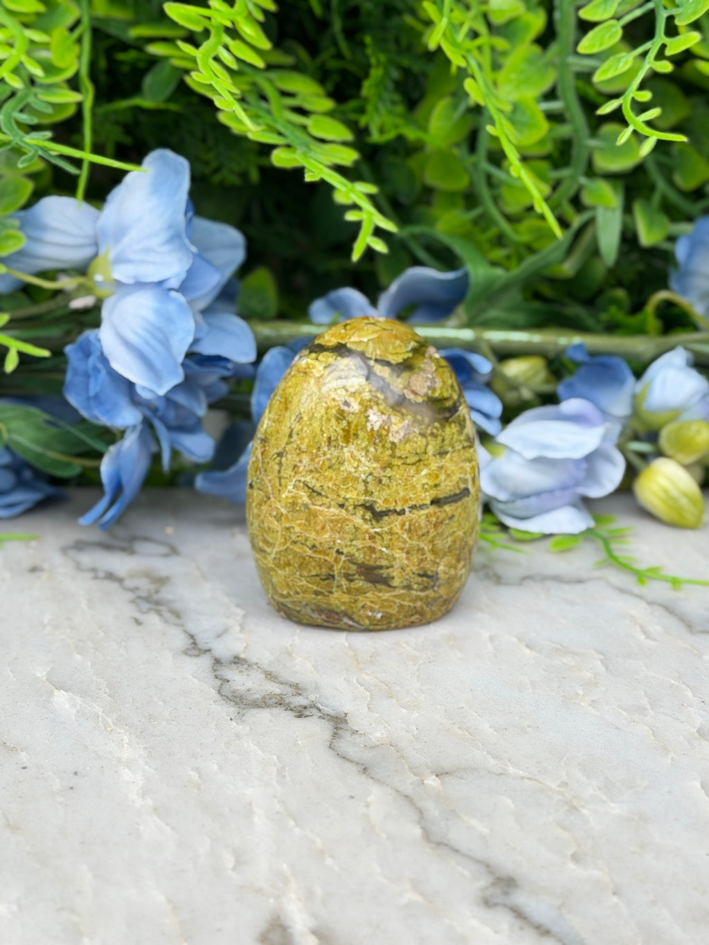 a yellowish-brown stone or rock placed on a marble surface, surrounded by blue flowers and green foliage.