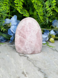 A pink crystal rests on a marble surface, surrounded by blue flowers and green foliage.