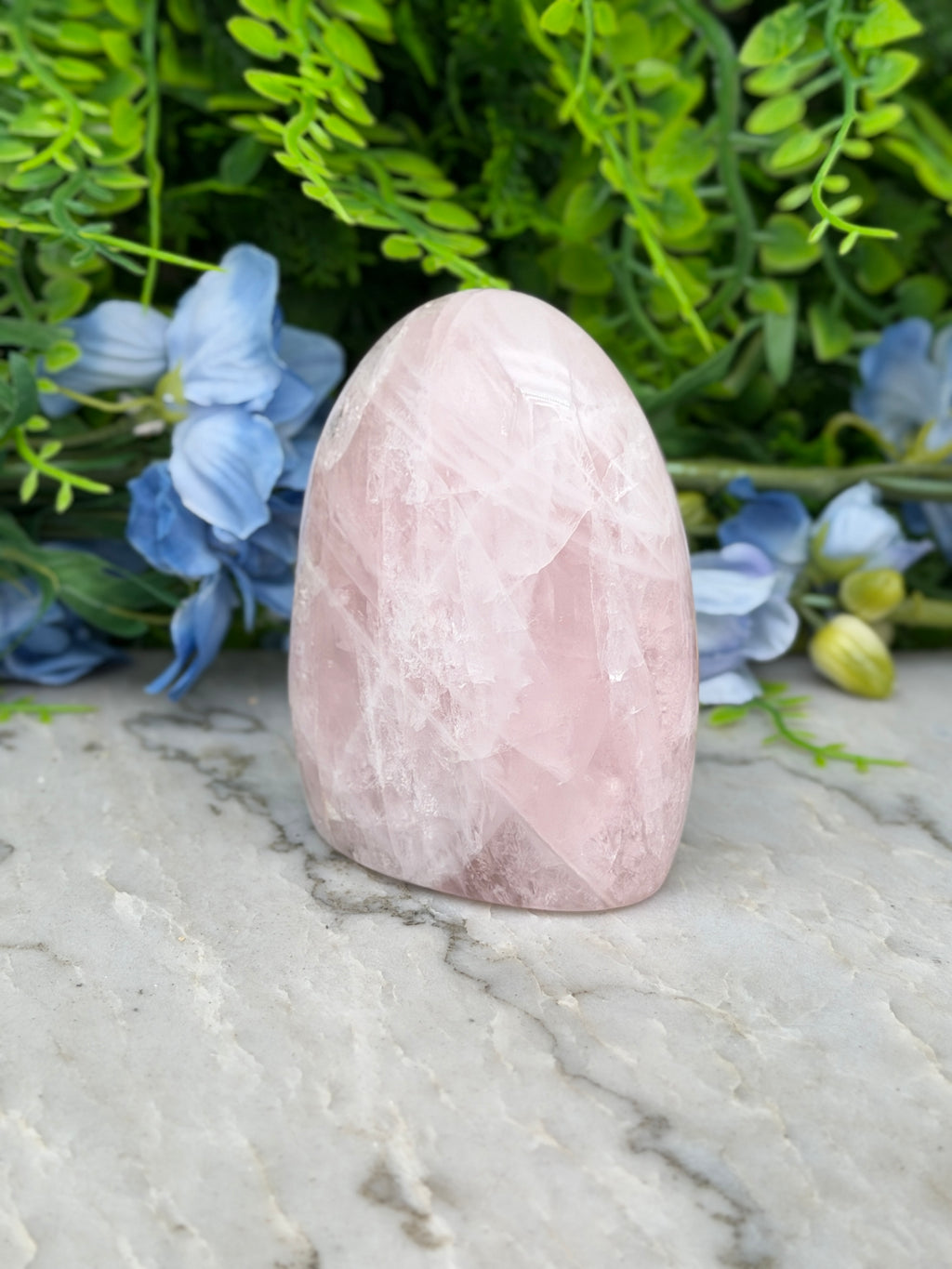 A pink crystal rests on a marble surface, surrounded by blue flowers and green foliage.
