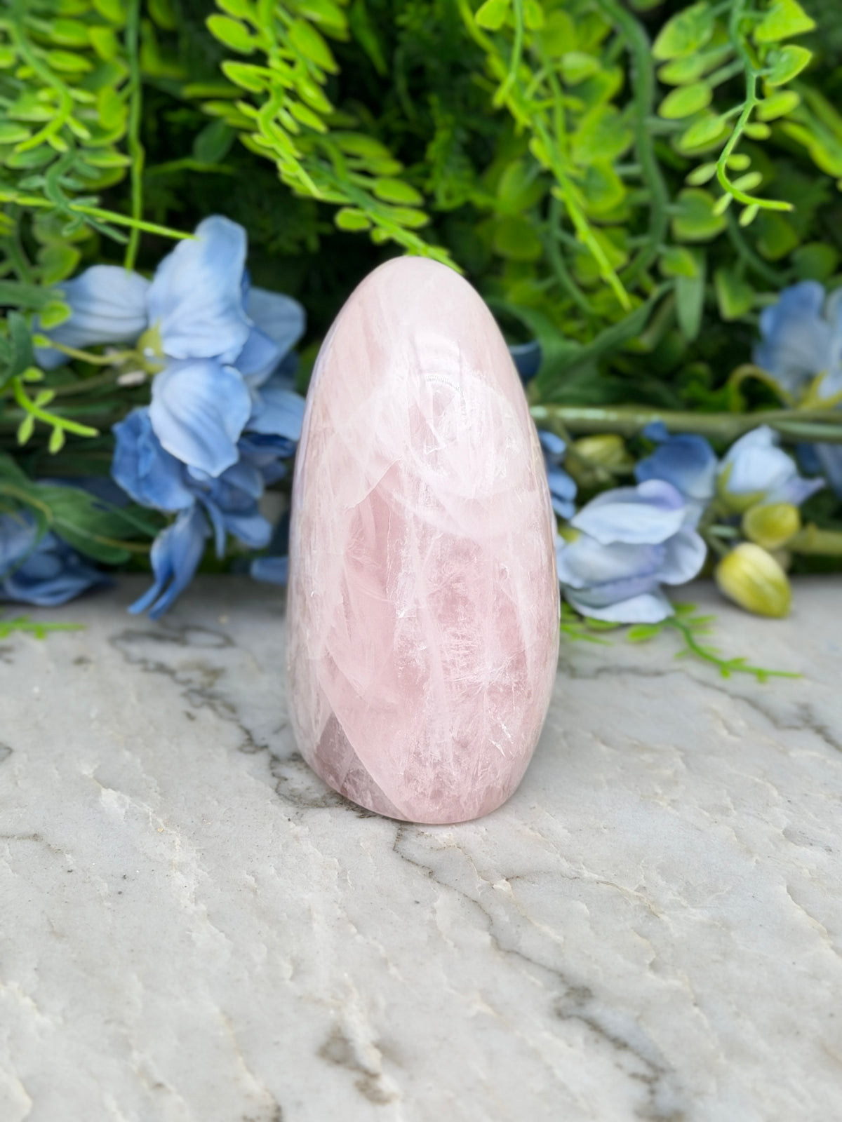 A pink crystal rests on a marble surface, surrounded by blue flowers and green foliage.