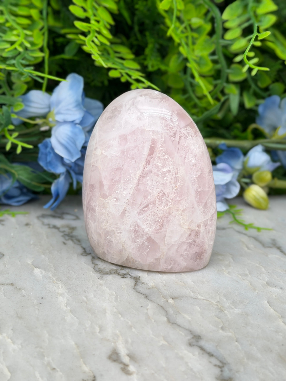A pink crystal rests on a marble surface, surrounded by lush green foliage and blue flowers.