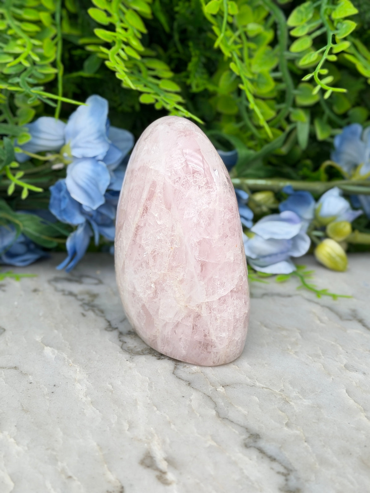 A pink crystal rests on a marble surface, surrounded by blue flowers and green foliage.