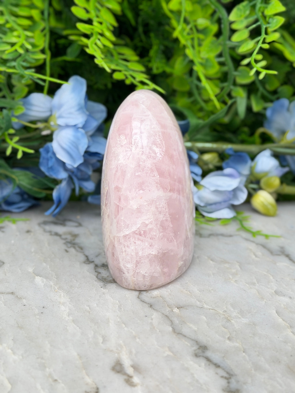A pink crystal rests on a marble surface, surrounded by blue flowers and greenery.
