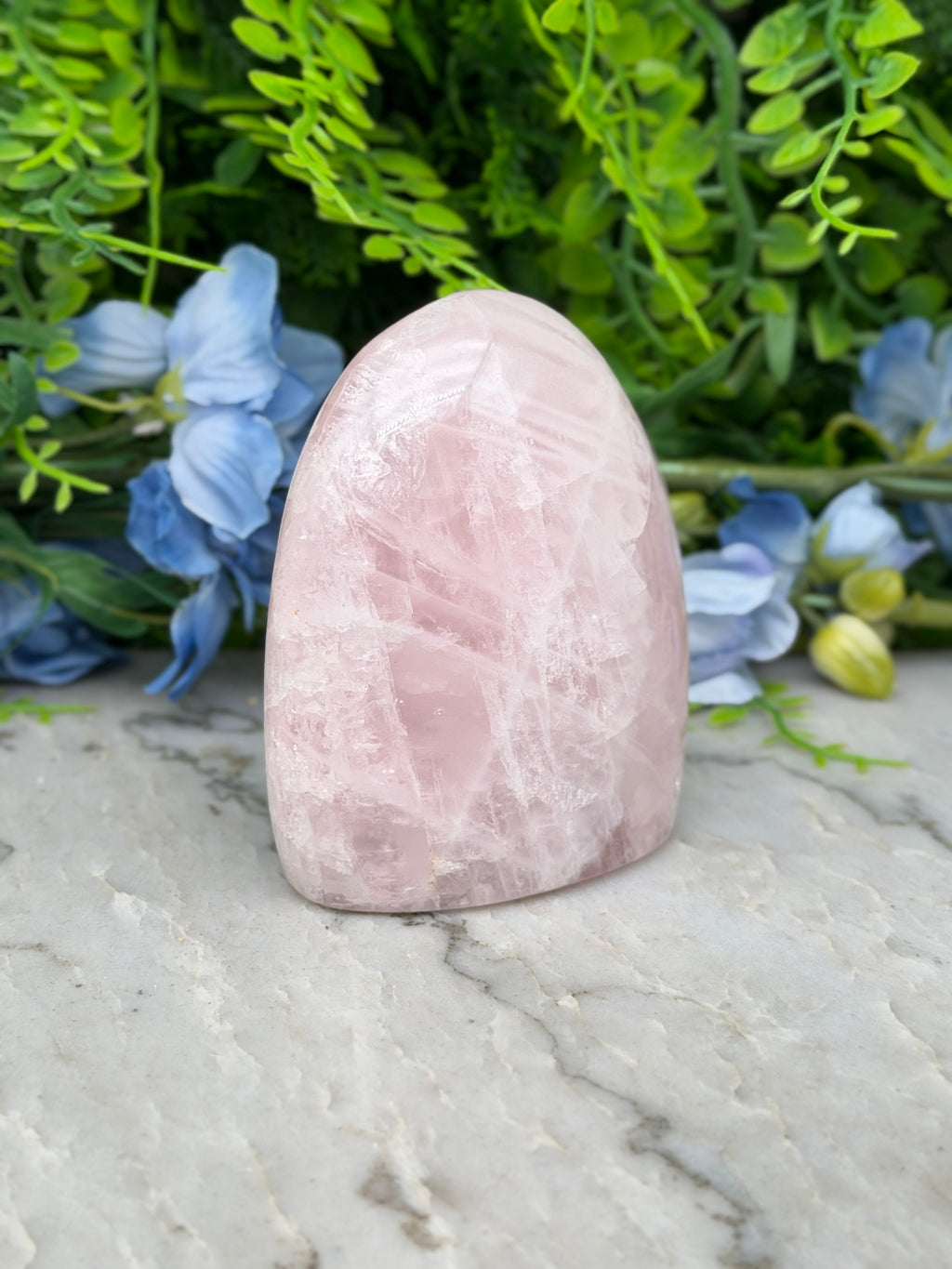 A pink crystal rests on a marble surface, surrounded by lush green foliage and blue flowers.