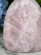 A close-up image of a pink crystal with a textured surface, placed on a marble surface next to a blue flower.