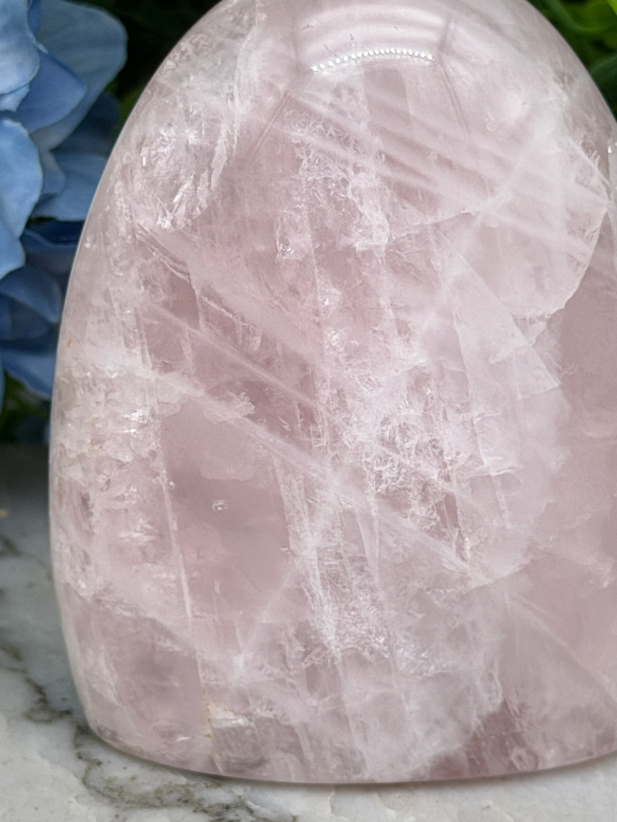 A close-up image of a pink crystal with a textured surface, placed on a marble surface next to a blue flower.