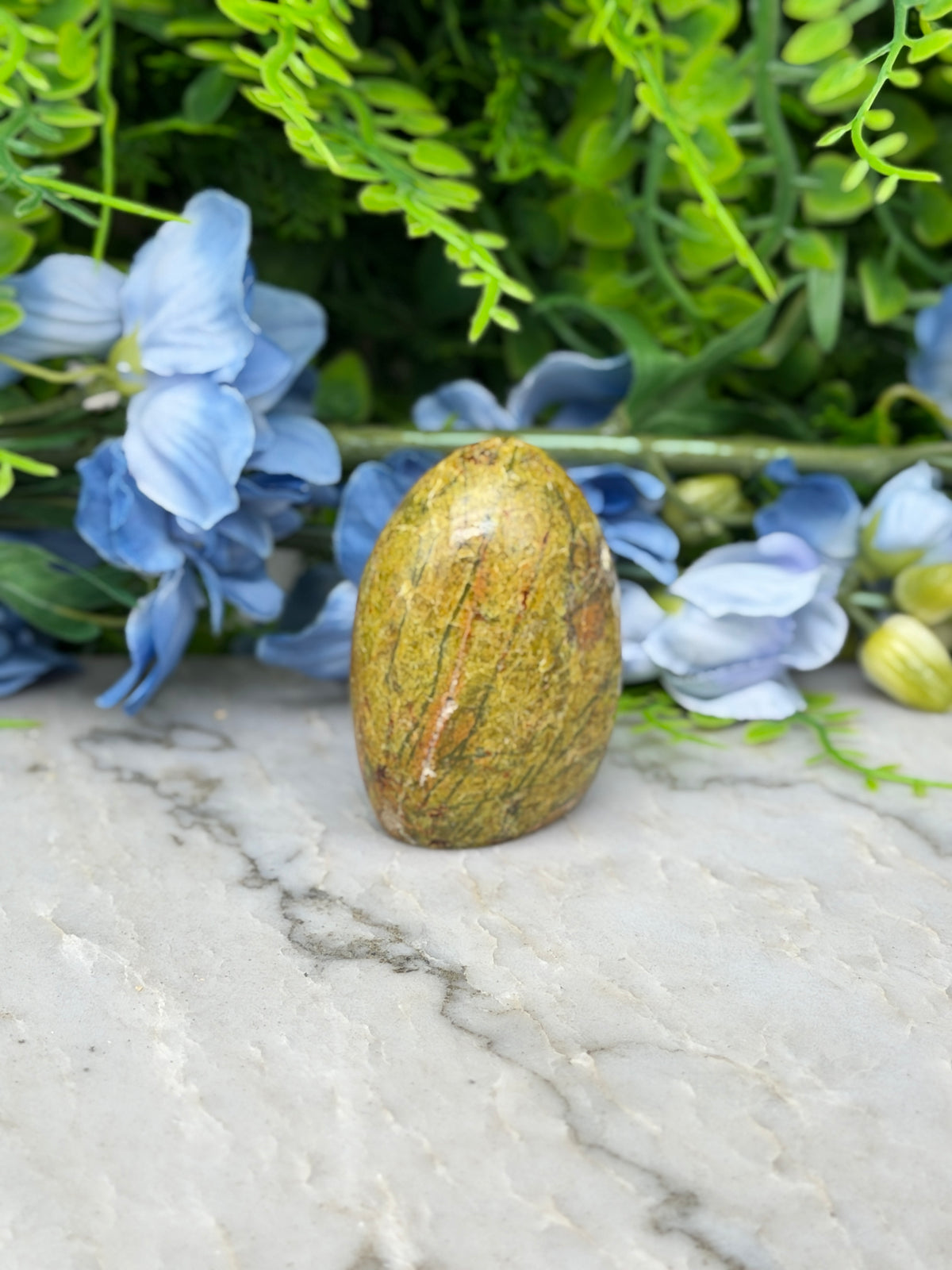 a yellowish-brown stone, possibly a jade or amber, surrounded by blue flowers and green foliage.