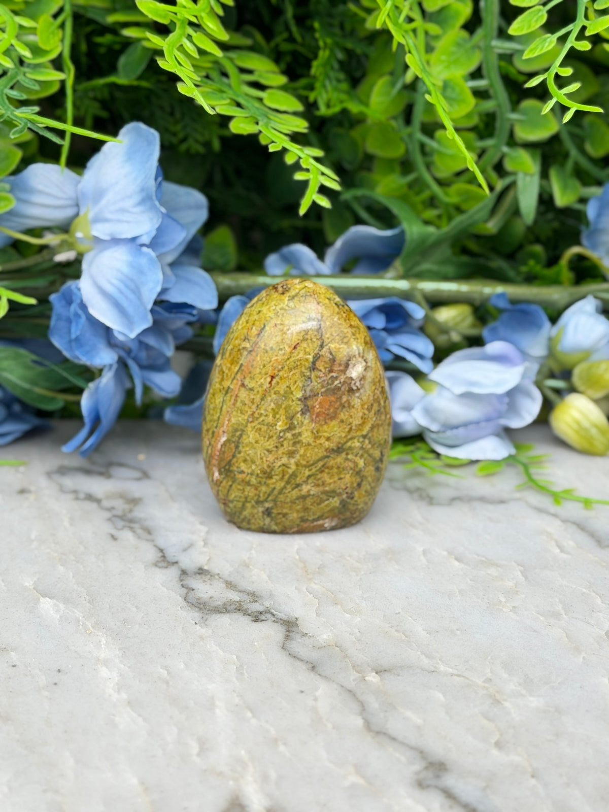 a yellowish-brown stone, possibly a jade egg, surrounded by blue flowers and green foliage.