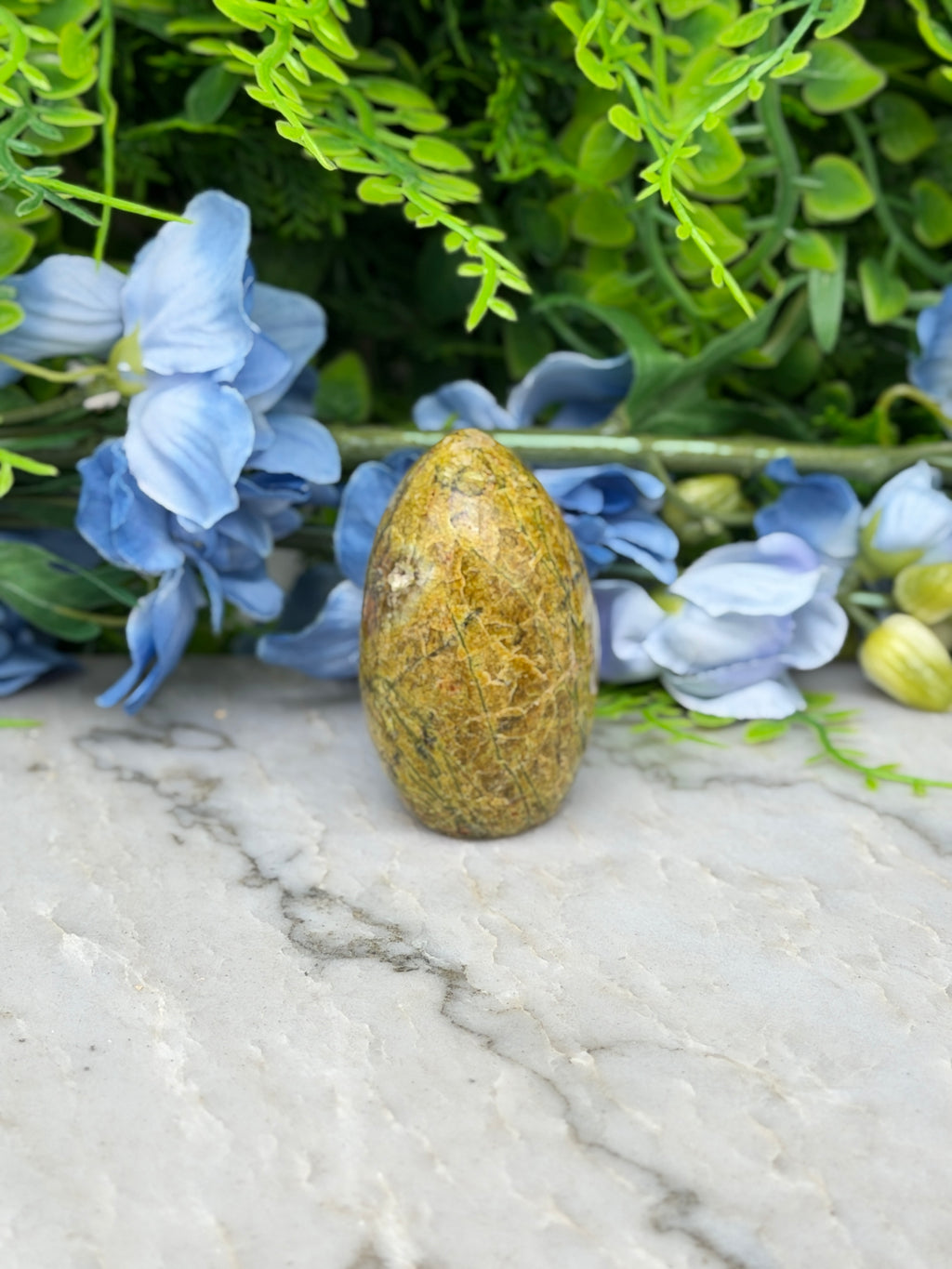a yellow egg-shaped object placed on a marble surface, surrounded by blue flowers and green foliage.