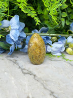 a yellow egg-shaped object placed on a marble surface, surrounded by blue flowers and green foliage.