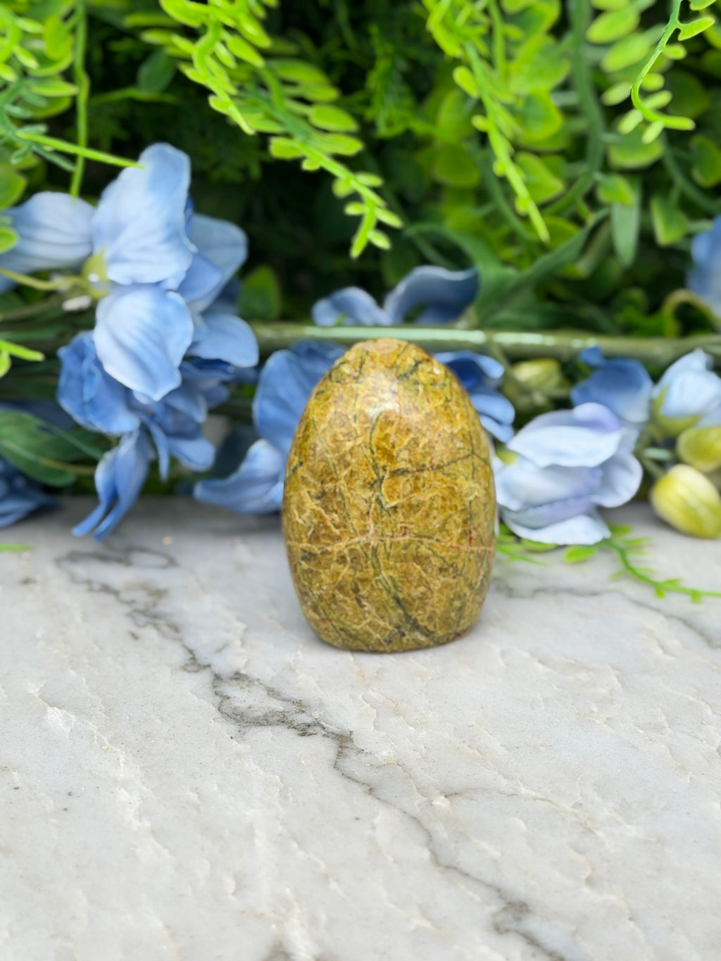 a yellowish-brown stone or gemstone placed on a marble surface, surrounded by blue flowers and greenery.