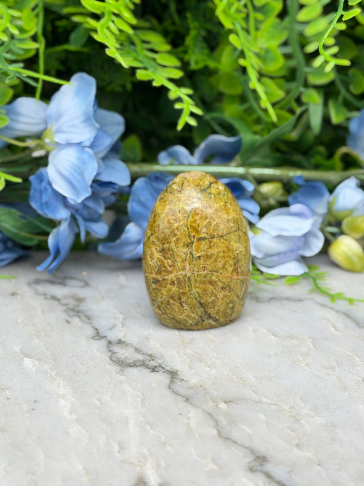 a yellowish-brown stone or gemstone placed on a marble surface, surrounded by blue flowers and greenery.