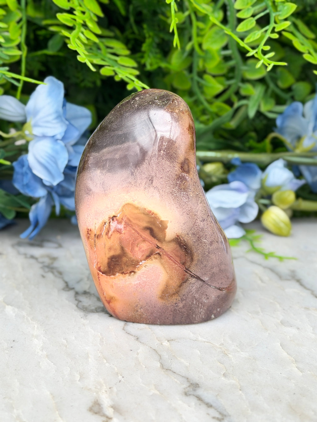 a brown rock with a leaf-like pattern on it, placed on a marble surface. The rock is surrounded by blue flowers, creating a natural and serene atmosphere.